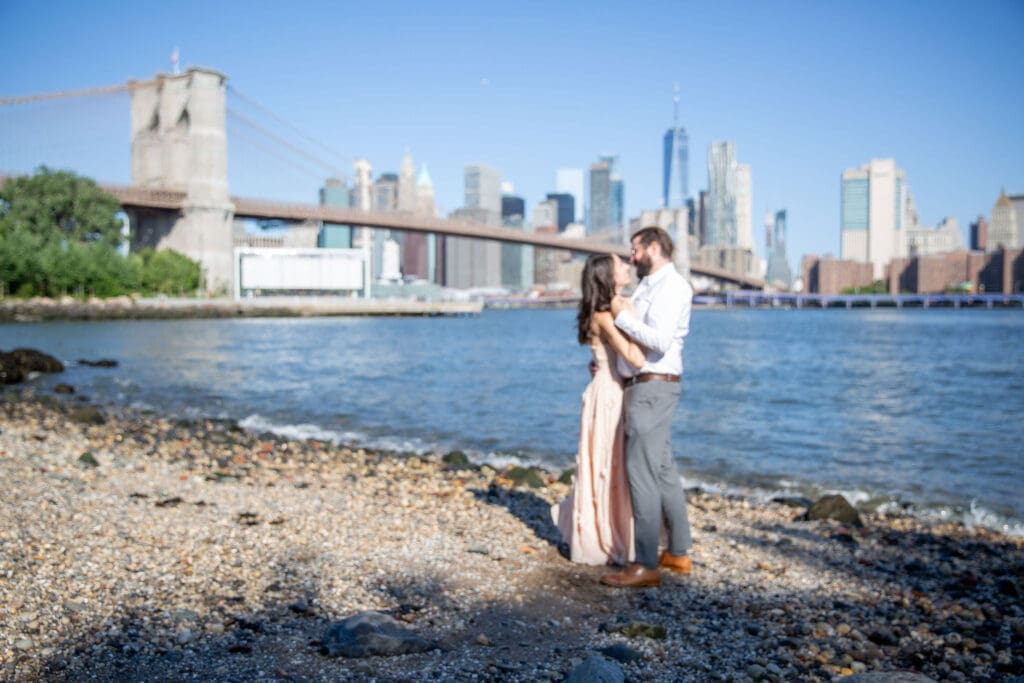 An engaged couple embrace and laugh in Pebble Beach in Dumbo, Brooklyn, NYC with the bridge and Manhattan skyline behind them.