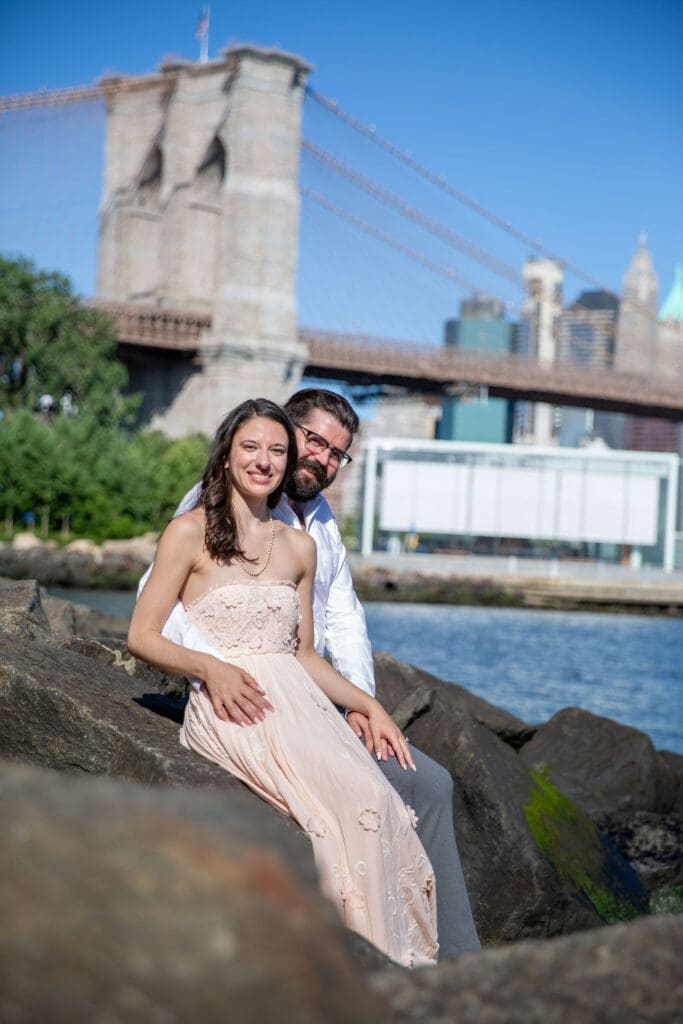 An engaged couple embrace and laugh in Brooklyn Bridge Park in Dumbo, Brooklyn, NYC with the bridge and Manhattan skyline behind them.