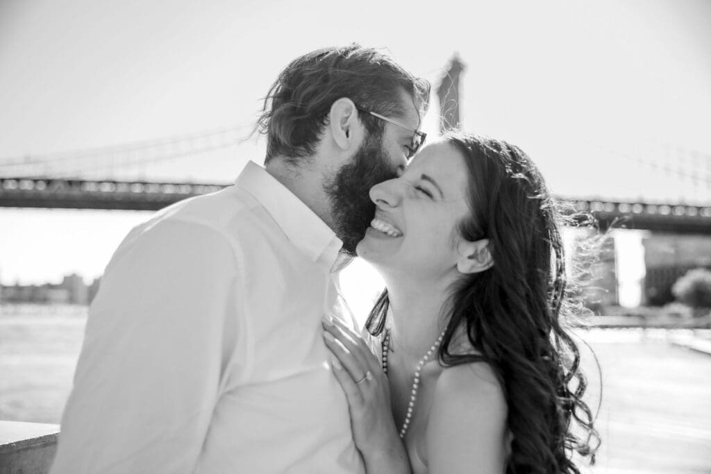 An engaged couple embrace and laugh in Brooklyn Bridge Park in Dumbo, Brooklyn, NYC.