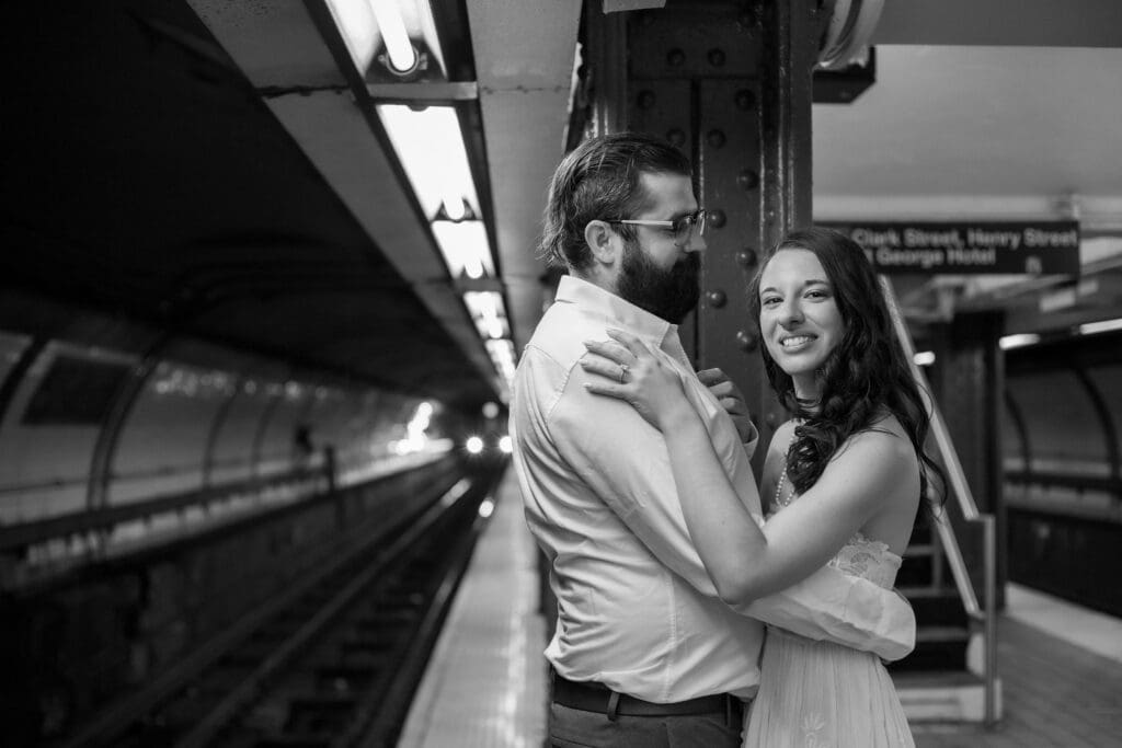 An engaged couple embraces on a Brooklyn subway platform as a train approaches.