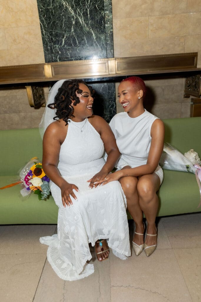 Two brides smile at the green couches after getting married at NYC City Hall.
