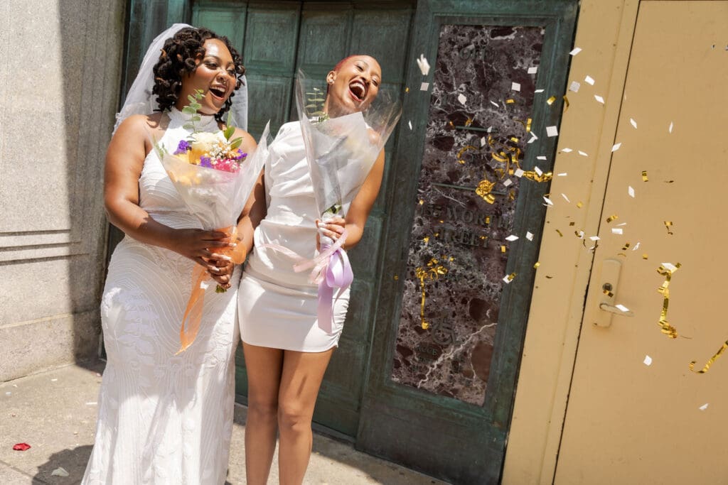 Two brides exit with confetti at NY City Hall's yellow doors and green wall after getting married.