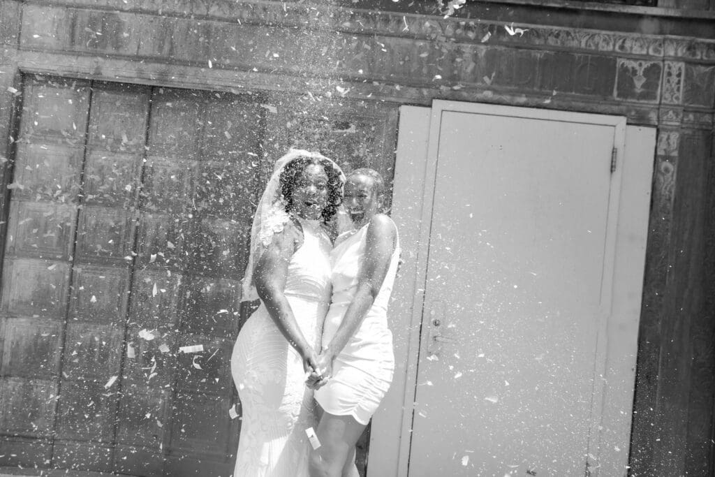 Two brides exit with confetti at NY City Hall's yellow doors and green wall after getting married. Black and White Image.