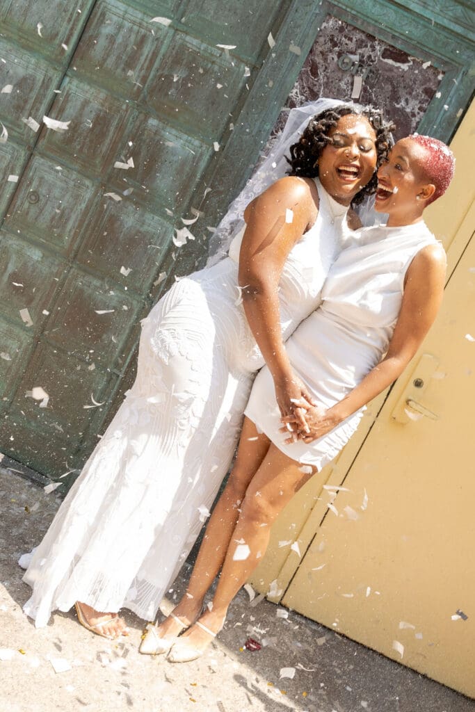Two brides exit with confetti at NY City Hall's yellow doors and green wall after getting married.