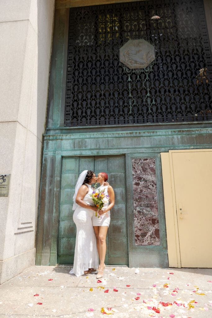 Two brides exit NY City Hall's yellow doors and green wall after getting married.
