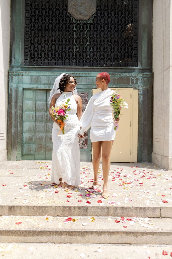 Two brides exit NY City Hall's yellow doors and green wall after getting married.