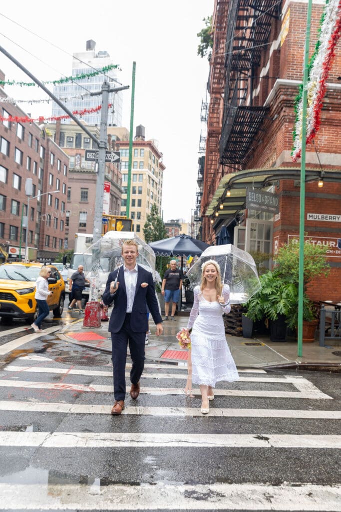Newlyweds take wedding portraits walking across the street with umbrellas in NYC's Little Italy.