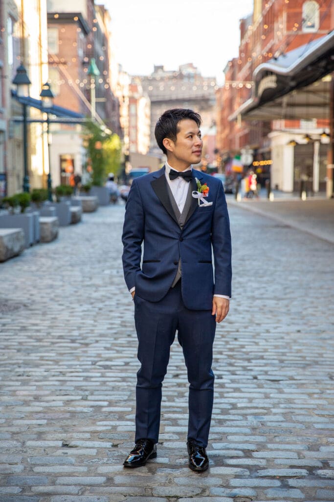 Groom in a blue suit poses for a solo photo with South Street Seaport in NYC behind him.