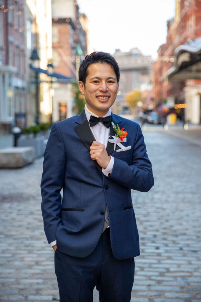 Groom in a blue suit poses for a solo photo with South Street Seaport in NYC behind him.