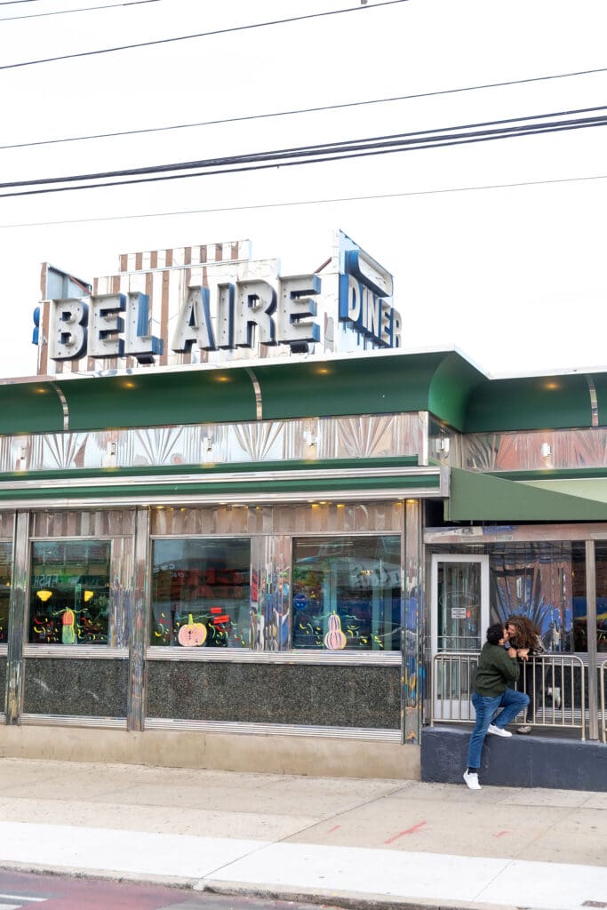 Engaged couple kiss in front of Bel-Aire Diner during their casual pre-wedding portrait session in Astoria, Queens.