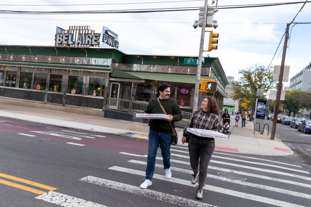 Engaged couple walk across the street from Bel-Aire Diner, holding pizza boxes during their casual pre-wedding portrait session in Astoria, Queens.