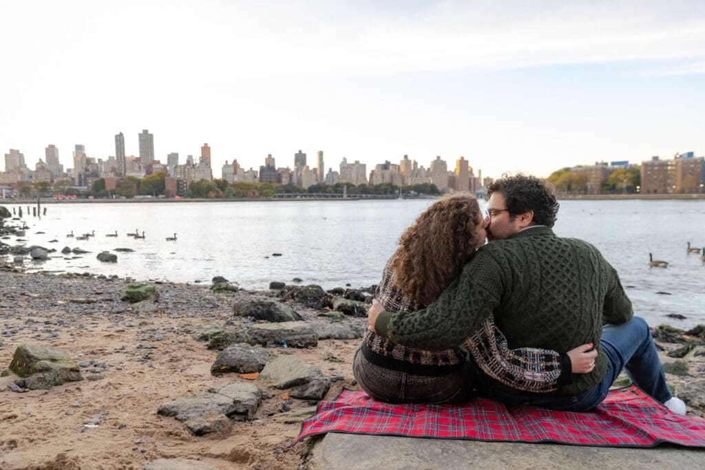 Engaged couple kiss at sunset at Hallett's Cove in Astoria, Queens for their fall engagement session. Behind them is the NYC skyline.
