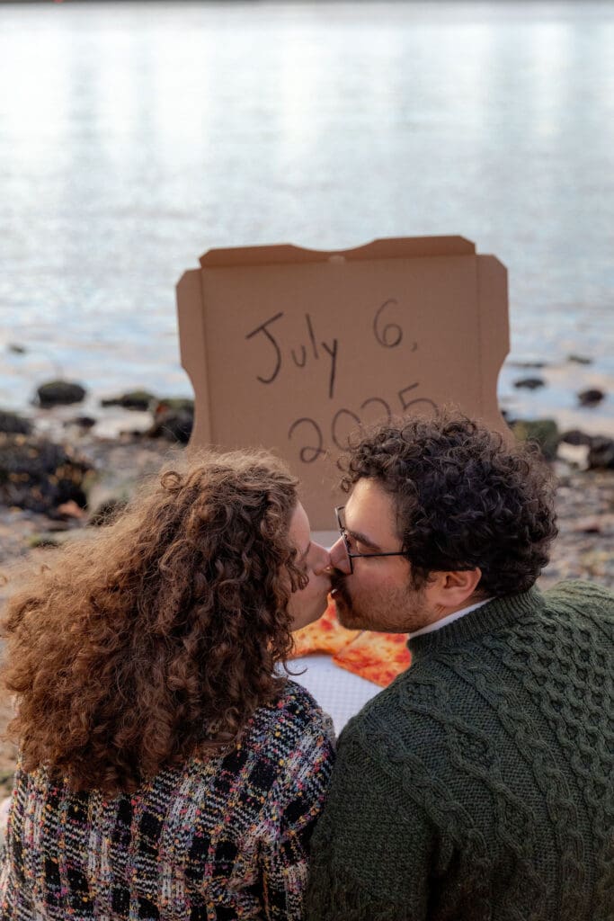 Engaged couple kiss at sunset at Hallett's Cove in Astoria, Queens for their fall engagement session. Behind them, a pizza box displays their wedding date and the NYC skyline.