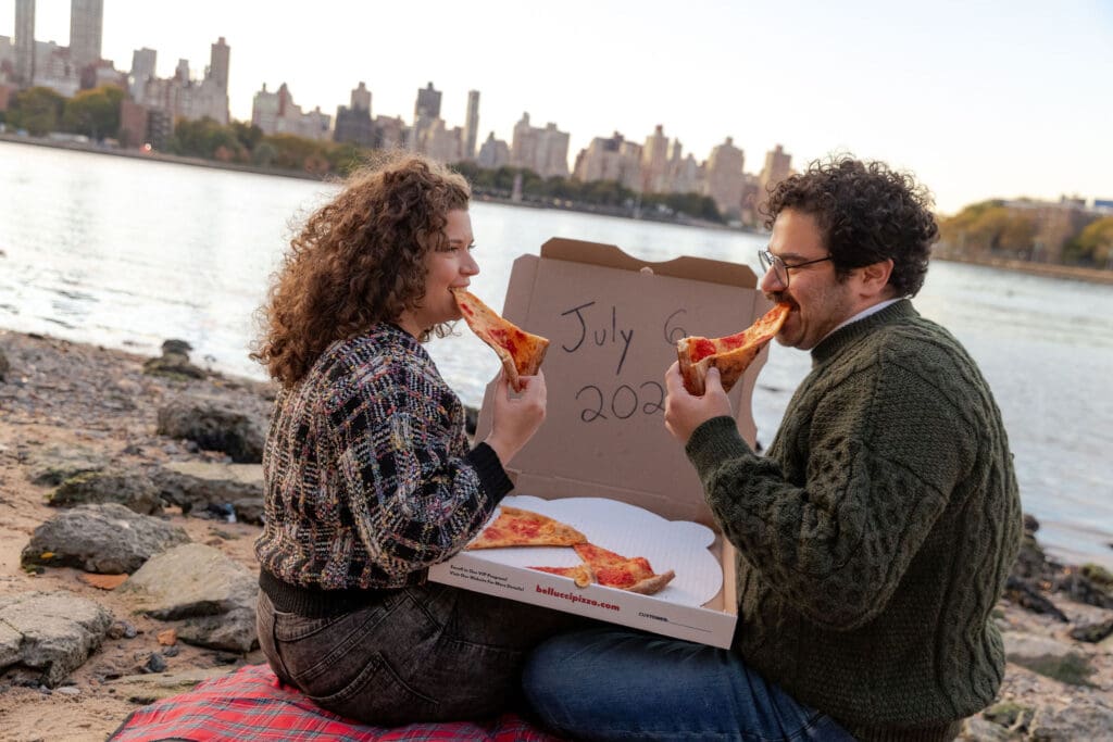 Engaged couple eat pizza at sunset at Hallett's Cove in Astoria, Queens for their fall engagement session. Behind them, a pizza box displays their wedding date and the NYC skyline.